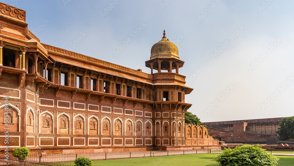 Jahangiri Mahal Palace. There are carvings, white ornaments, windows on the sandstone walls. A tower with a dome on a blue sky background. Arched entrance.  A green lawn nearby. India. Agra.