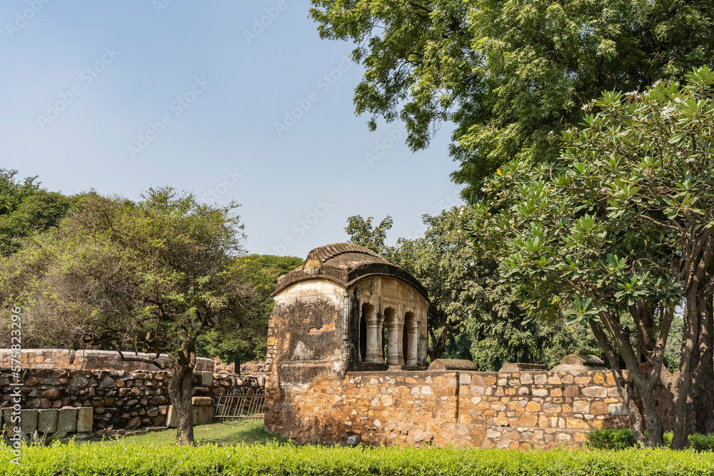 Ruins of an ancient building in the Qutub Minar complex. Weathered cracked walls, columns, arched roof. There is lush green vegetation around. Blue sky. India. Delhi
