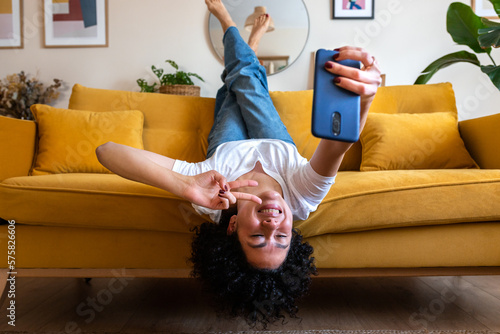 Joyful African American woman lying upside down on the sofa takes selfie with smart phone. Social media.