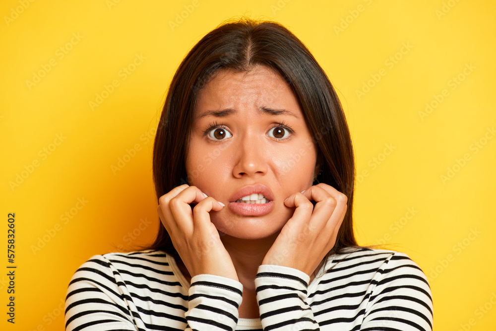 Close up shot of scared Asian brunette woman stares bugged eyes keeps hands near mouth has puzzled expression dressed in lined jumper poses against yellow background Close up shot of scared Asian brunette woman stares bugged eyes keeps hands near mouth has puzzled expression dressed in lined jumper poses against yellow background