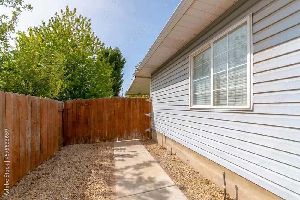 Wooden fence gate at the side of the house with gray wood lap siding and paned windows. Concrete pathway heading to the side-hinged gate against the trees and sky at the back. Wooden fence gate at the side of the house with gray wood lap siding and paned windows. Concrete pathway heading to the side-hinged gate against the trees and sky at the back.