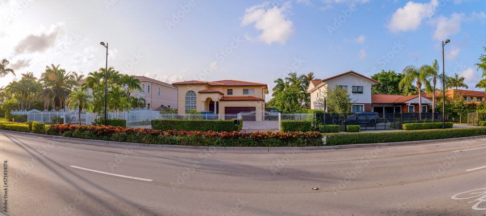 Miami, Florida- Panorama of gated mediterranean houses against the sunny sky. There is a road at the front with bushes and street lights at the front of the houses with gates and fences.