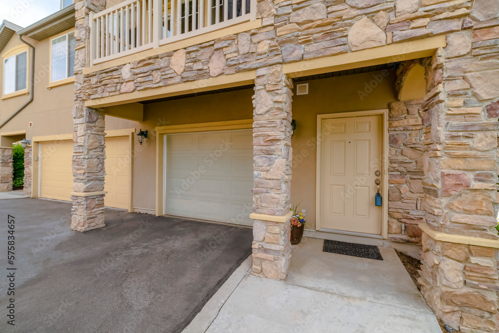 House entrance with attached double garages under the balcony with stone veneer walls. There is a cream door with lockbox on the right near the white garage door beside the cream garage door.