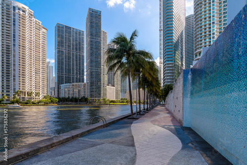 Miami River Walk with wall and coconut trees against the high-rise buildings at Miami, Florida. Concrete path near the river on the right with views of modern multi-storey buildings. © Jason