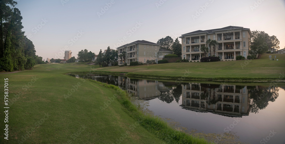 Panorama of apartment buildings with lakefront view against sunset sky in Destin, Florida. Lake with green field shore and reflections of the buildings on its water surface.