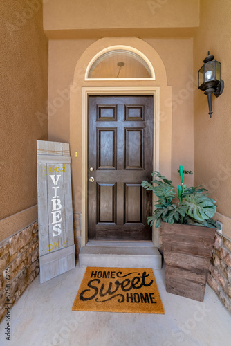 Vertical shot of a black front door with decorative wood sign stand and potted plants. There is an arched transom window above the door beside the antique wall lamp on the right.