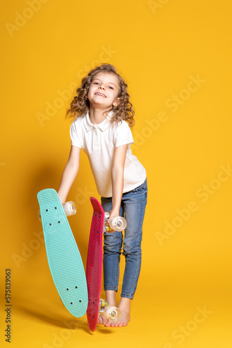 Full length of little girl in white polo, blue jeans who holds two skateboards on yellow background