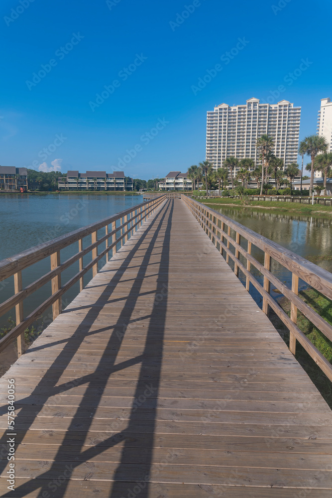 Obraz premium Vertical shot of boardwalk over the water with views of buildings at Destin, Florida. Wooden boardwalk with railings over the lake against the apartment buildings and blue sky background.