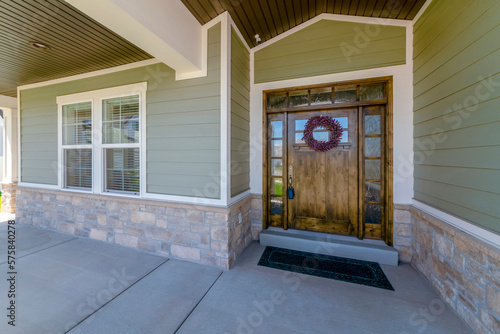 Wide wood front door with glass panes, wreath, and lockbox of a house. House exterior with green wood lap and stone veneer siding under a ceiling with security camera at the door near the windows.