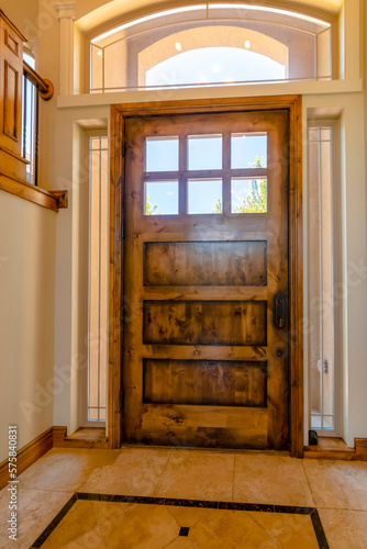 Interior view of a house with closed glass paned brown wooden front door. A transom window, deadbolt, and the sunny outdoors can be seen from the foyer.