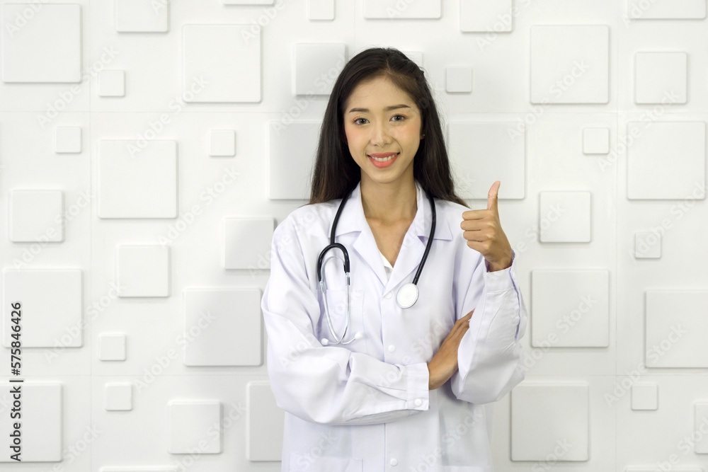 Young asian doctor in white gown and stethoscope stand with finger thumb up in front of white rectangle pattern ceramic wall. Young asian doctor in white gown and stethoscope stand with finger thumb up in front of white rectangle pattern ceramic wall.