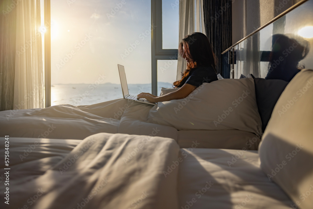 Happy fresh relaxed asian woman resting sitting on bed in hotel room with two beds using laptop enjoying good morning after wakeup in cozy room with soft light from window. Happy fresh relaxed asian woman resting sitting on bed in hotel room with two beds using laptop enjoying good morning after wakeup in cozy room with soft light from window.