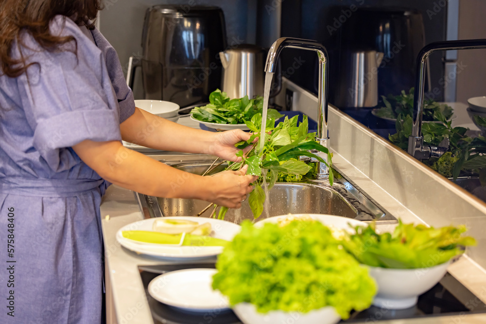 Woman wearing bathrobe washing a fresh Thai basil and other fresh vegetable to remove pesticides before cooking in kitchen under the tap.