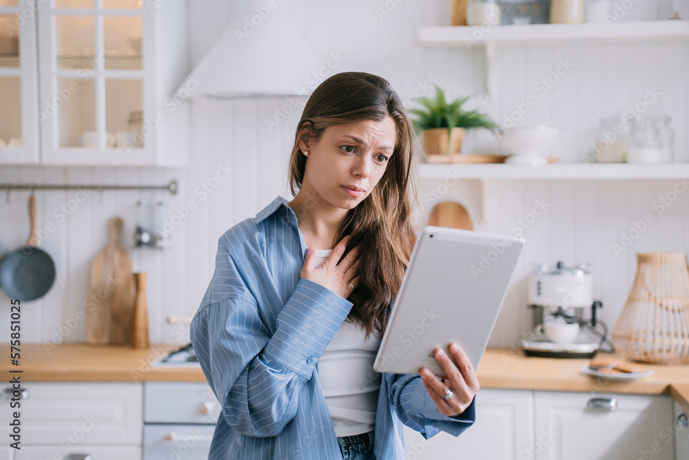 Confused pretty caucasian woman makes video call using tablet touches chest feels frustration unhappy by received news. Dark haired Italian housewife in blue shirt talks with parents at kitchen. Grief Confused pretty caucasian woman makes video call using tablet touches chest feels frustration unhappy by received news. Dark haired Italian housewife in blue shirt talks with parents at kitchen. Grief