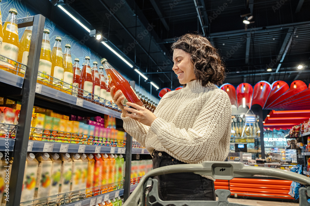 Shopper selects juice in grocery store with cart - obrazy, fototapety, plakaty 