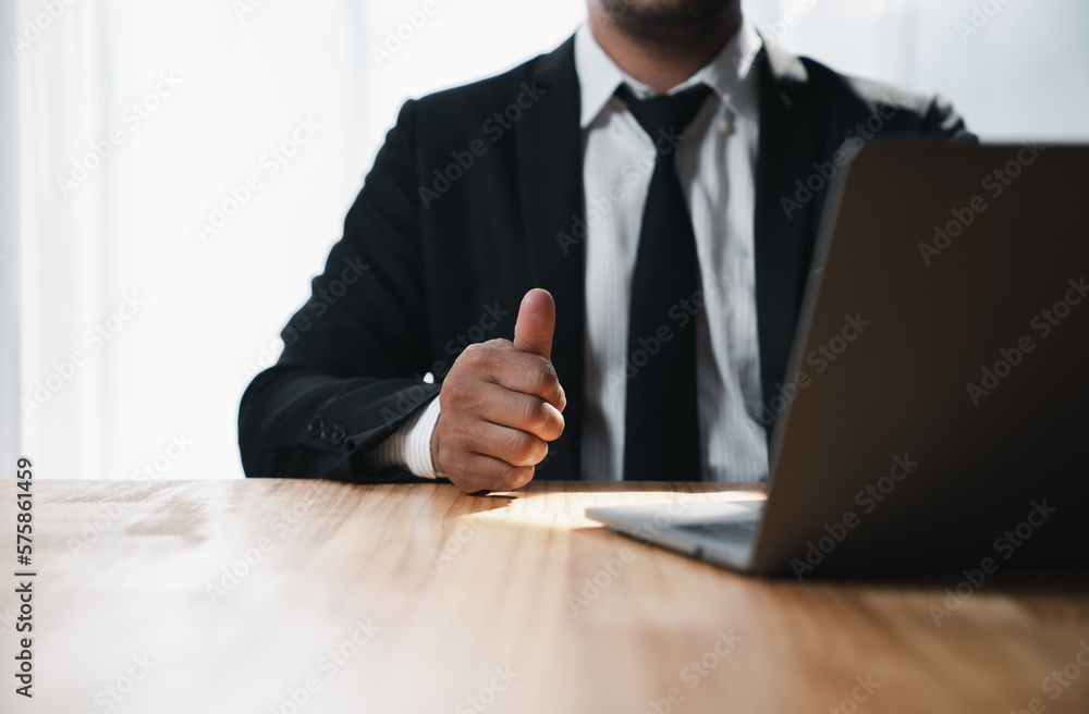 A young Asian businessman in a black suit giving great thumbs up to real estate business success looking at a laptop screen in the office room. A young Asian businessman in a black suit giving great thumbs up to real estate business success looking at a laptop screen in the office room.
