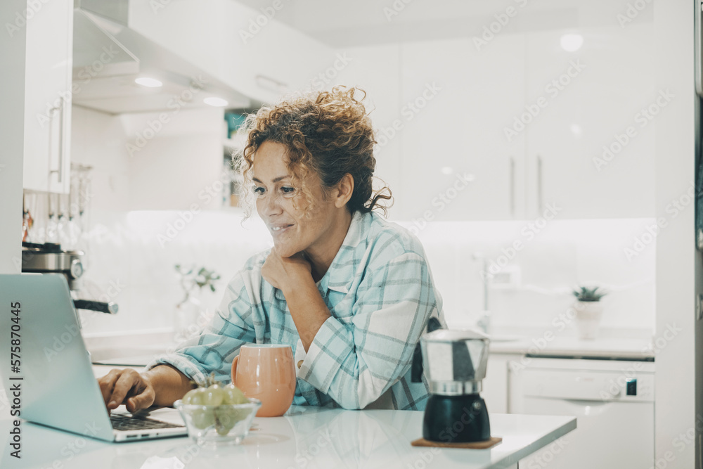 One happy cheerful woman smile in the kitchen doing morning breakfast and reading notifications or new e-mail on a laptop computer. White ambient house and modern online worker female people alone One happy cheerful woman smile in the kitchen doing morning breakfast and reading notifications or new e-mail on a laptop computer. White ambient house and modern online worker female people alone