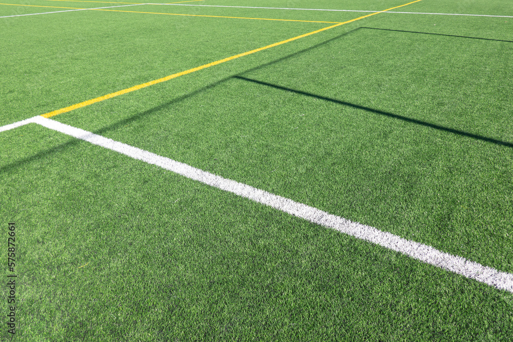 Obraz premium White and yellow stripes on green soccer field from side view. Artificial turf on football field. Green synthetic grass on sport ground with shadow from soccer goal net