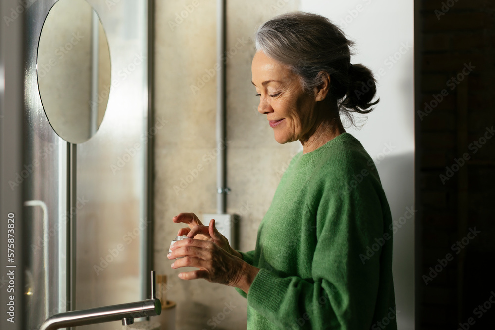 Smiling woman with moisturizer standing in front of mirror at home Smiling woman with moisturizer standing in front of mirror at home