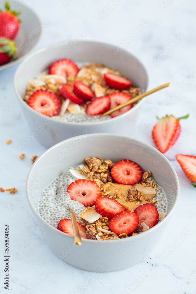 Chia pudding with homemade coconut granola, peanut butter and strawberries in a gray bowl, marble background. Healthy diet, detox, summer recipe.
