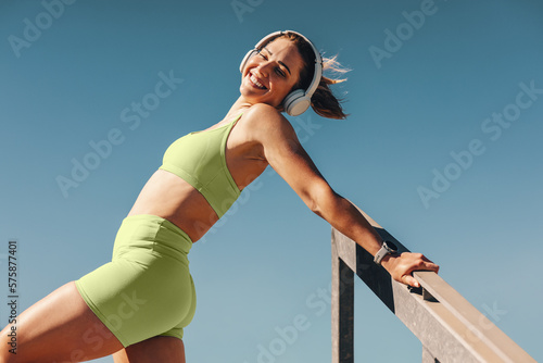 Fototapeta Naklejka Na Ścianę i Meble -  Fit and healthy woman smiling while listening to music with headphones and stretching her body outdoors