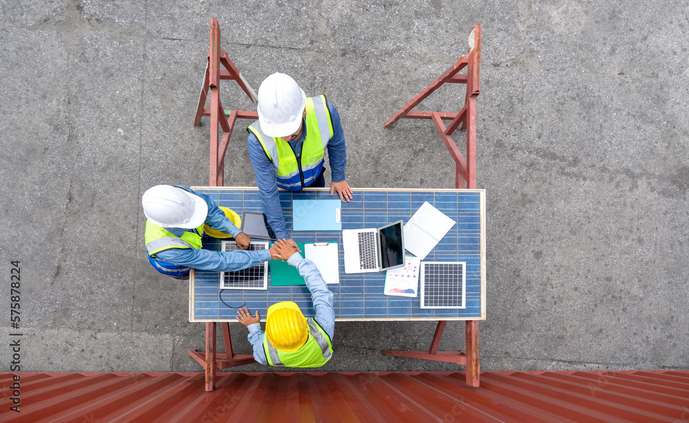 Group of shipment worker in hardhat and safety vest .join hands together on a table made of large solar cell panel. A large container is in the background. Top View