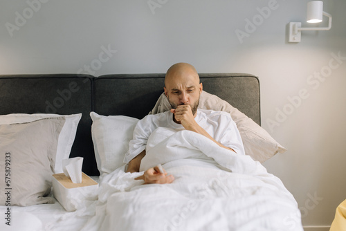 A man lying in bed looking tired and sickly. He covers his mouth with his hand when he coughs, and in his other hand he holds a thermometer with a pack of dry tissues next to him.