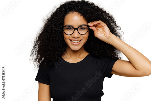 Lovely young African American woman grins at camera, keeps hand on rim of spectacles, glad to hear good news, dressed in casual black t shirt, isolated over brown background. Happiness concept