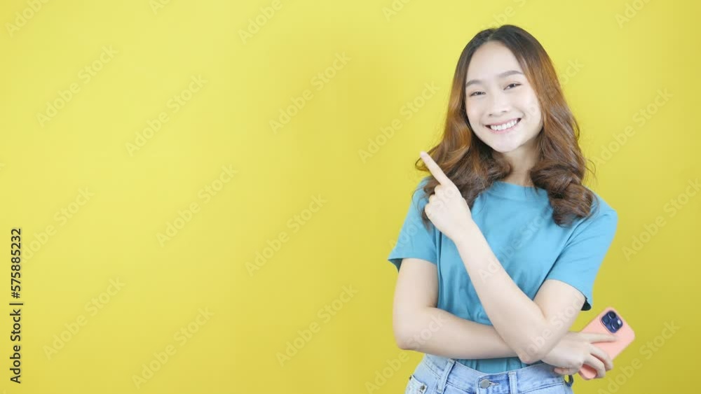 Video of Asian woman smiling while pointing left and thumbs up introducing product standing on yellow background. Video of Asian woman smiling while pointing left and thumbs up introducing product standing on yellow background.