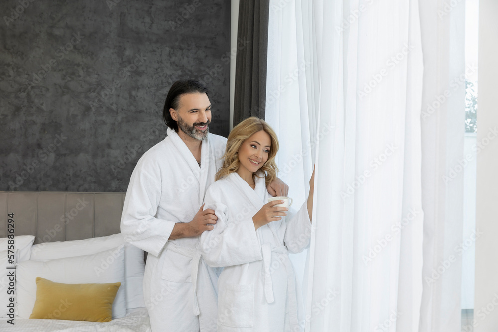 Handsome couple in bathrobe in the bedroom standing near the window curtains Handsome couple in bathrobe in the bedroom standing near the window curtains
