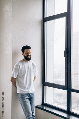 A guy with a beard in white T-shirts and blue jeans stands by the window in a new apartment and rejoices in happiness.