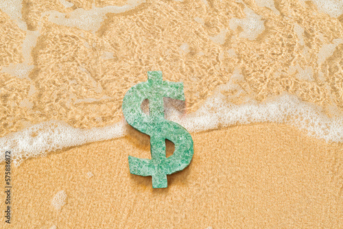 A stone in the form of a dollar sign lies on a sandy beach. The currency is flooded with surf water. Background on the theme of hotel business and recreation at the resort.