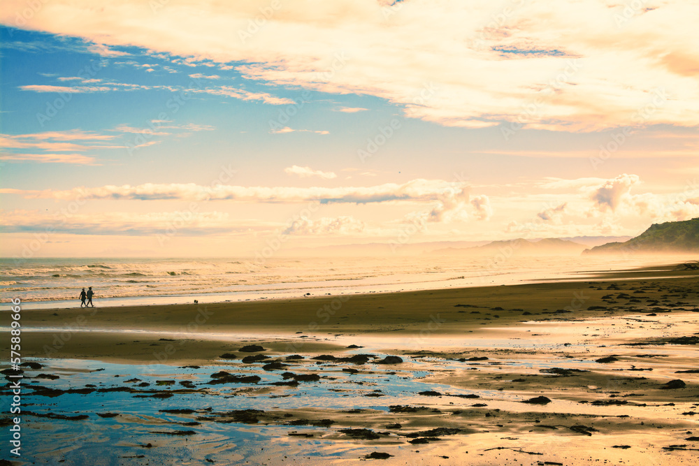 Naklejka premium Active couple walking their pet dog along a vast sand beach at sunset. Pacific Ocean coastline near Gisborne, New Zealand