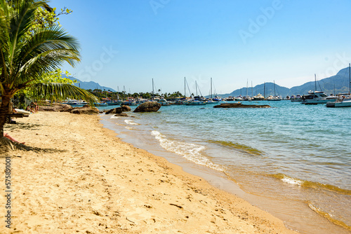 View of a deserted beach, footprints on the yellow sand, palm trees and boats parked in the marina in the background. Ilhabela, Sao Paulo