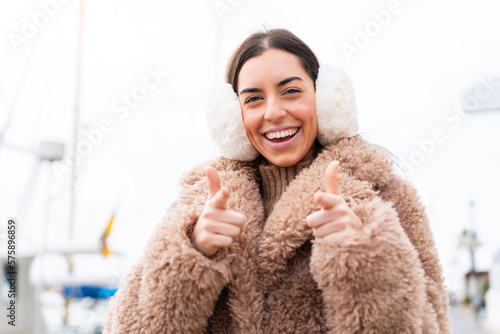 Young woman wearing winter muffs at outdoors pointing to the front and smiling photo