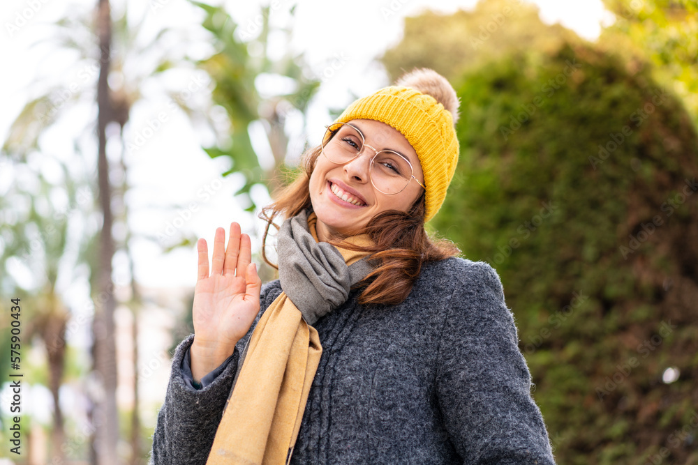 Brunette woman wearing winter jacket at outdoors saluting with hand with happy expression