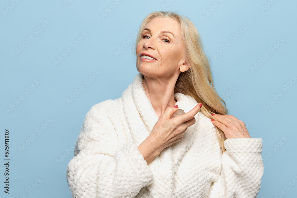 Getting ready. Beautiful middle-aged woman in bathrobe applying perfumes over blue studio background. Concept of natural beauty, face skin care, cosmetology and cosmetics, health Getting ready. Beautiful middle-aged woman in bathrobe applying perfumes over blue studio background. Concept of natural beauty, face skin care, cosmetology and cosmetics, health