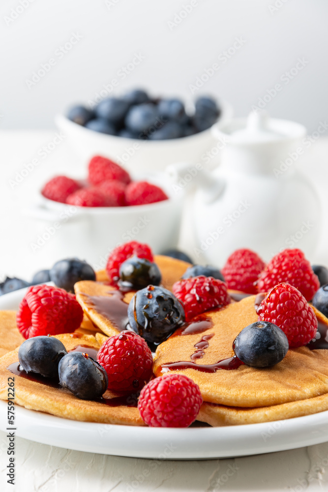 Close-up of pancakes with caramel on plate with blueberries and raspberries with bowls and jar on white table, selective focus, vertical Close-up of pancakes with caramel on plate with blueberries and raspberries with bowls and jar on white table, selective focus, vertical