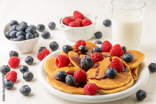 Top view of pancakes with caramel on plate with blueberries and raspberries with bowls, glass of milk and jug on white table, selective focus, horizontal