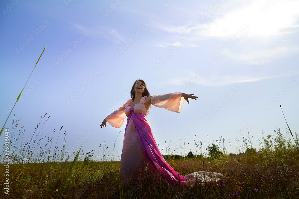 Beautiful girl in a lush pink ball gown in green field during blooming of flowers and blue sky on background. Model posing on nature landscape as princess from fary tale Beautiful girl in a lush pink ball gown in green field during blooming of flowers and blue sky on background. Model posing on nature landscape as princess from fary tale