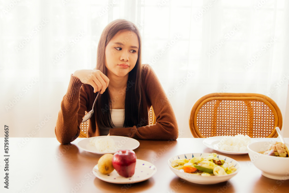 Young asian woman hungry but bored eating on table full of bland food sitting looking at food with boredom and having anorexia, not happy to eat and not having good appetite. Young asian woman hungry but bored eating on table full of bland food sitting looking at food with boredom and having anorexia, not happy to eat and not having good appetite.