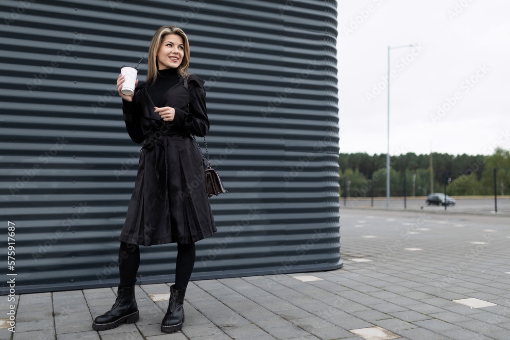 successful adult caucasian woman with a cup of coffee stands in the city near a gray embossed wall successful adult caucasian woman with a cup of coffee stands in the city near a gray embossed wall