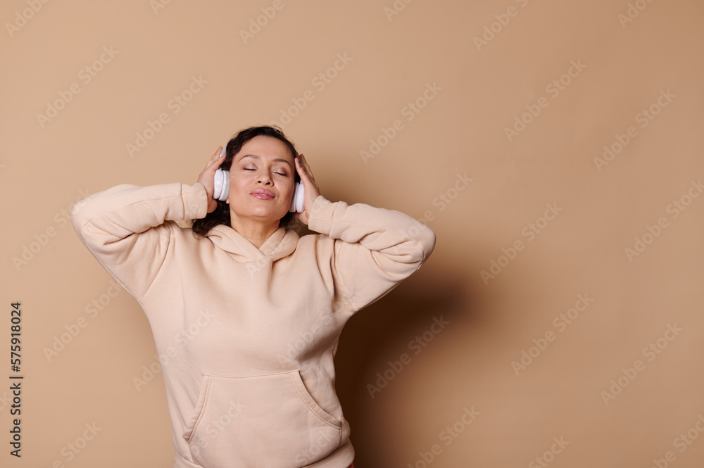 Latin American middle-aged attractive delightful pretty woman wearing wireless headphones and beige hoodie, enjoying soothing music on isolated cream background. People Entertainment Lifestyle Leisure Latin American middle-aged attractive delightful pretty woman wearing wireless headphones and beige hoodie, enjoying soothing music on isolated cream background. People Entertainment Lifestyle Leisure