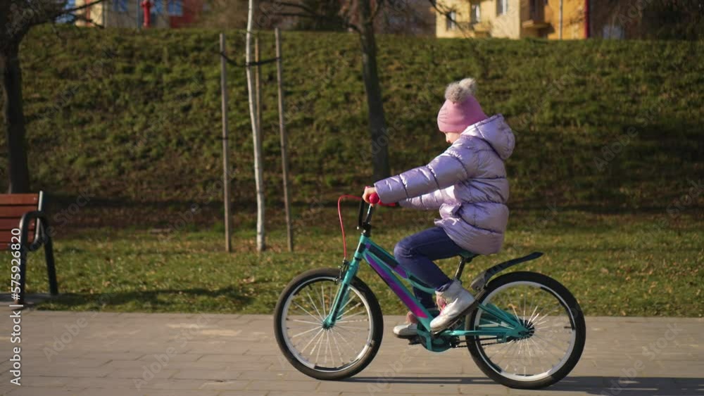 Girl riding bicycle in sunny winter in city park at beautiful sunset in slow motion. Happy childhood, sports outdoors in playground Girl riding bicycle in sunny winter in city park at beautiful sunset in slow motion. Happy childhood, sports outdoors in playground