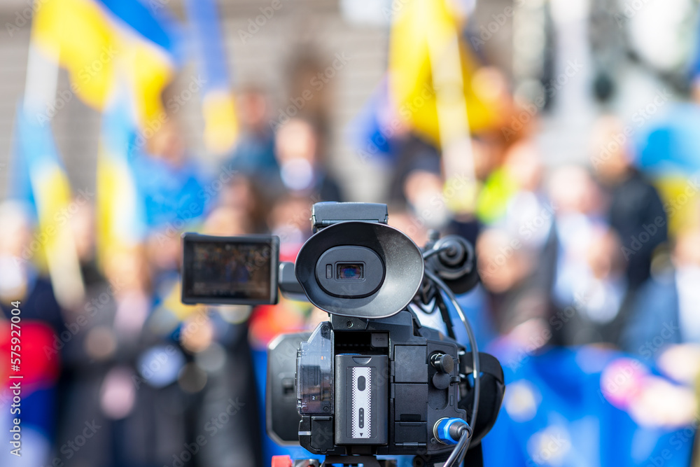 Anti-war meeting, focus on television video camera, blurred crowd of people with Ukrainian flags in the background Anti-war meeting, focus on television video camera, blurred crowd of people with Ukrainian flags in the background