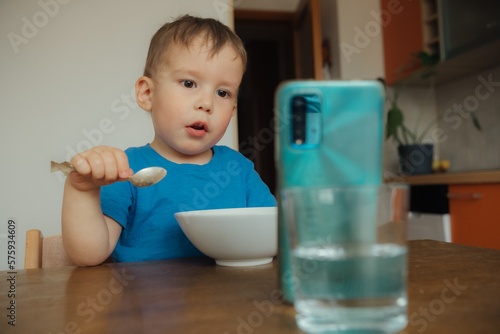  Child, a boy of three years old, is having breakfast at the kitchen table and watching cartoons on his smartphone