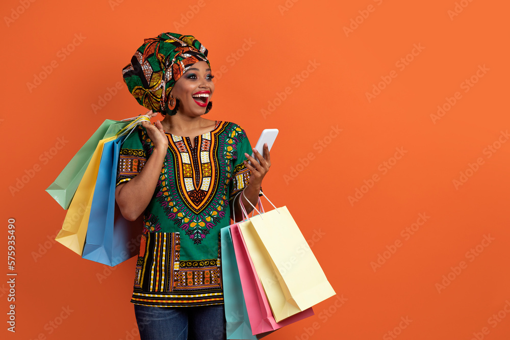 Excited traditional african woman shopping online, using smartphone on orange Excited traditional african woman shopping online, using smartphone on orange