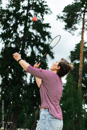 Outdoors against the forest and the sky on a cloudy cool day, a woman plays badminton, hits a red shuttlecock with a racket, copy space.