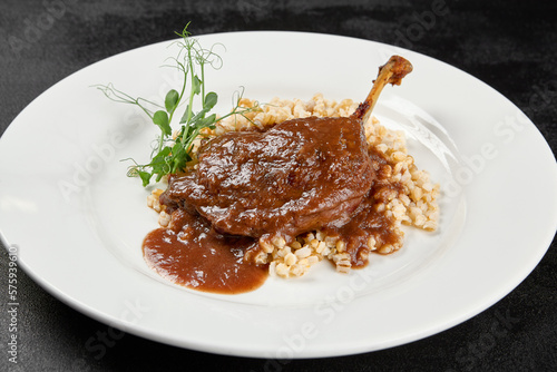 Classic dish french cuisine - roasted duck leg in porto sauce with garnish on black background. Cooked duck leg with pearl barley on white plate on dark concrete table. Duck confit in minimal style.