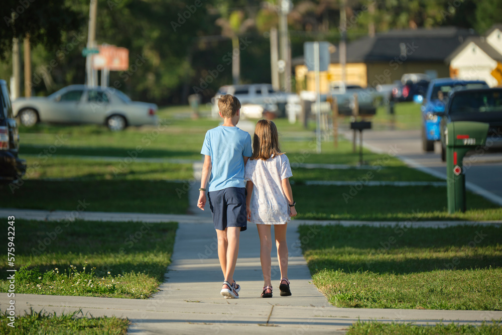 Rear view of two young teenage children, girl and boy, brother and sister walking together on rural street on bright sunny day. Vacation time concept Rear view of two young teenage children, girl and boy, brother and sister walking together on rural street on bright sunny day. Vacation time concept